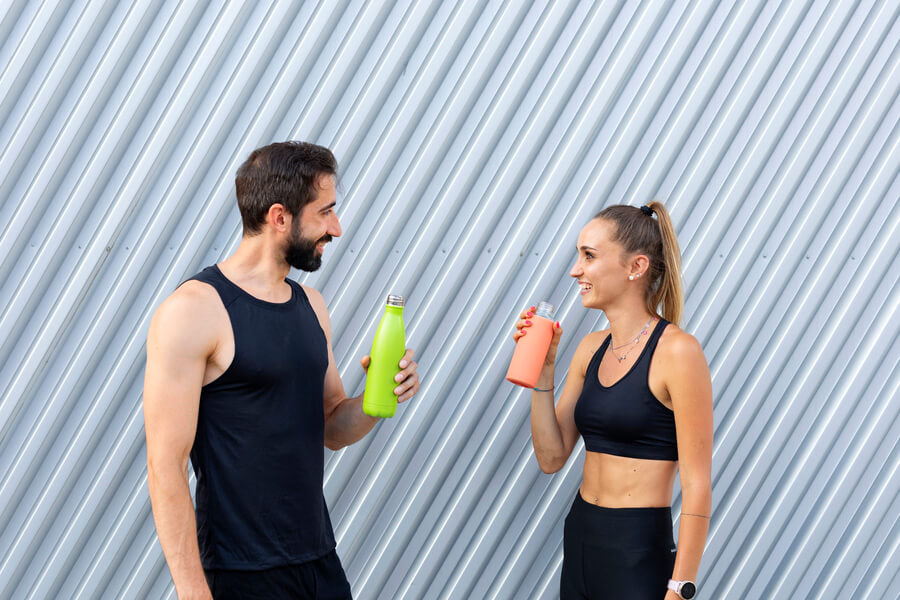 Hombre y mujer deportistas conversando mientras sostienen botellas de colores. - Como empezar a tomar suplementos deportivos