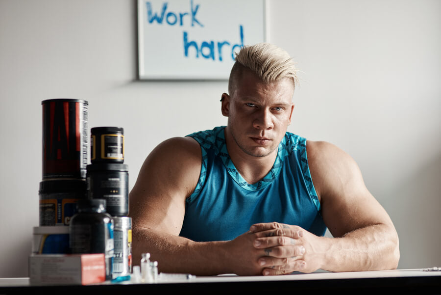 Como funcionan los suplementos deportivos - Hombre musculoso con camiseta azul sentado junto a suplementos deportivos en una mesa.