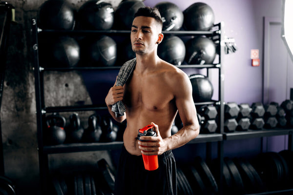  creatina monohidratada beneficios - Hombre sin camisa en gimnasio sosteniendo un batido post-entrenamiento.