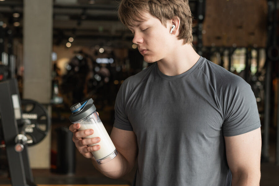 Hombre en gimnasio revisando suplemento proteico en botella.