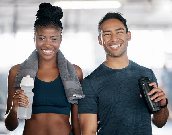 Dos personas sonrientes con ropa deportiva y botellas de agua en un gimnasio.