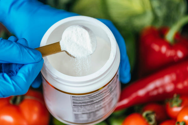 Polvo blanco en suplemento alimenticio siendo medido con cuchara sobre fondo de verduras frescas