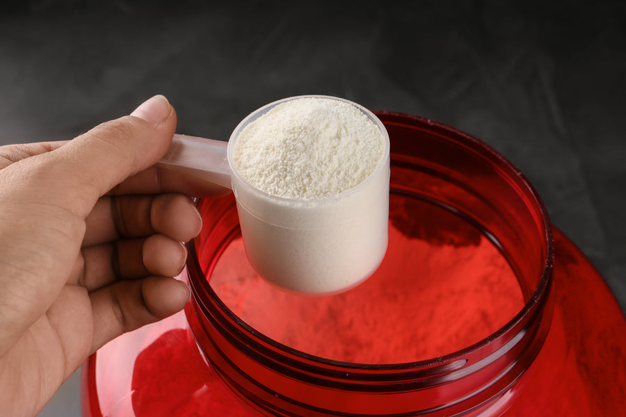 Mano sosteniendo scoop de proteína en polvo sobre envase rojo abierto