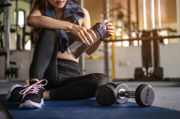 Proteína isolate gourmet - Mujer deportista sentada hidratándose junto a mancuernas.