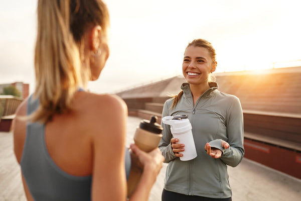Suplementos deportivos para mujeres - Dos mujeres deportistas conversando al aire libre con batidos proteicos al atardecer.