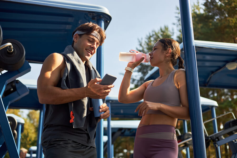 suplementos deportivos que funcionan - Hombre y mujer haciendo una pausa en el gimnasio al aire libre mientras revisan un celular.