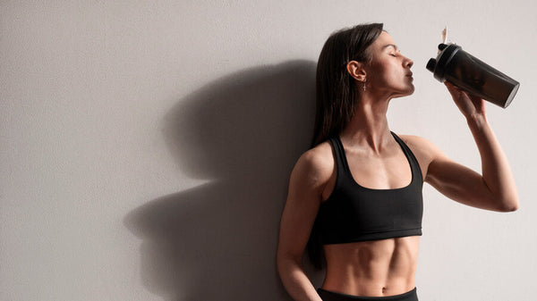 Suplementos deportivos tipos - Mujer deportista bebiendo de un shaker mientras descansa apoyada en la pared.